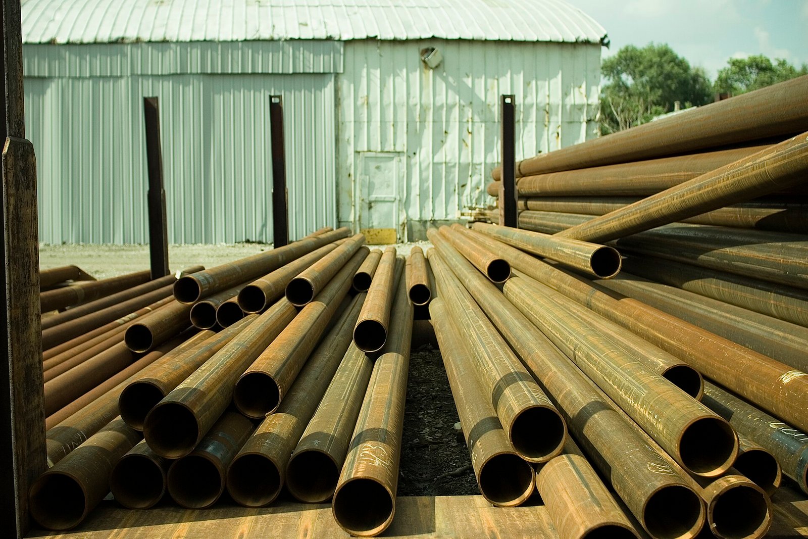 Round metal tubes with open circular ends and naturally weathered surfaces, stacked in long lengths.